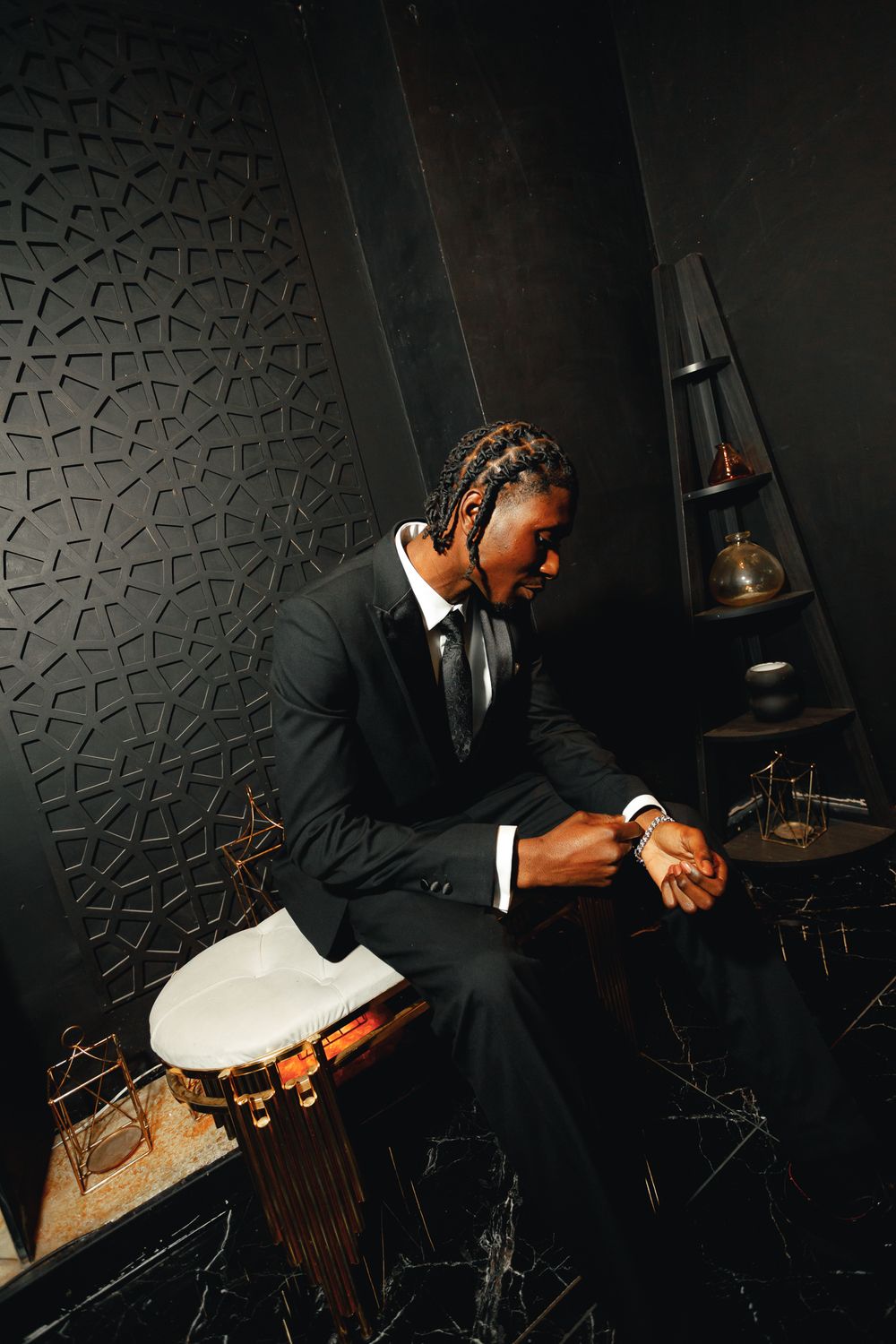 A musician in a black suit sits in a dark studio space with textured walls and drum equipment visible in the background.