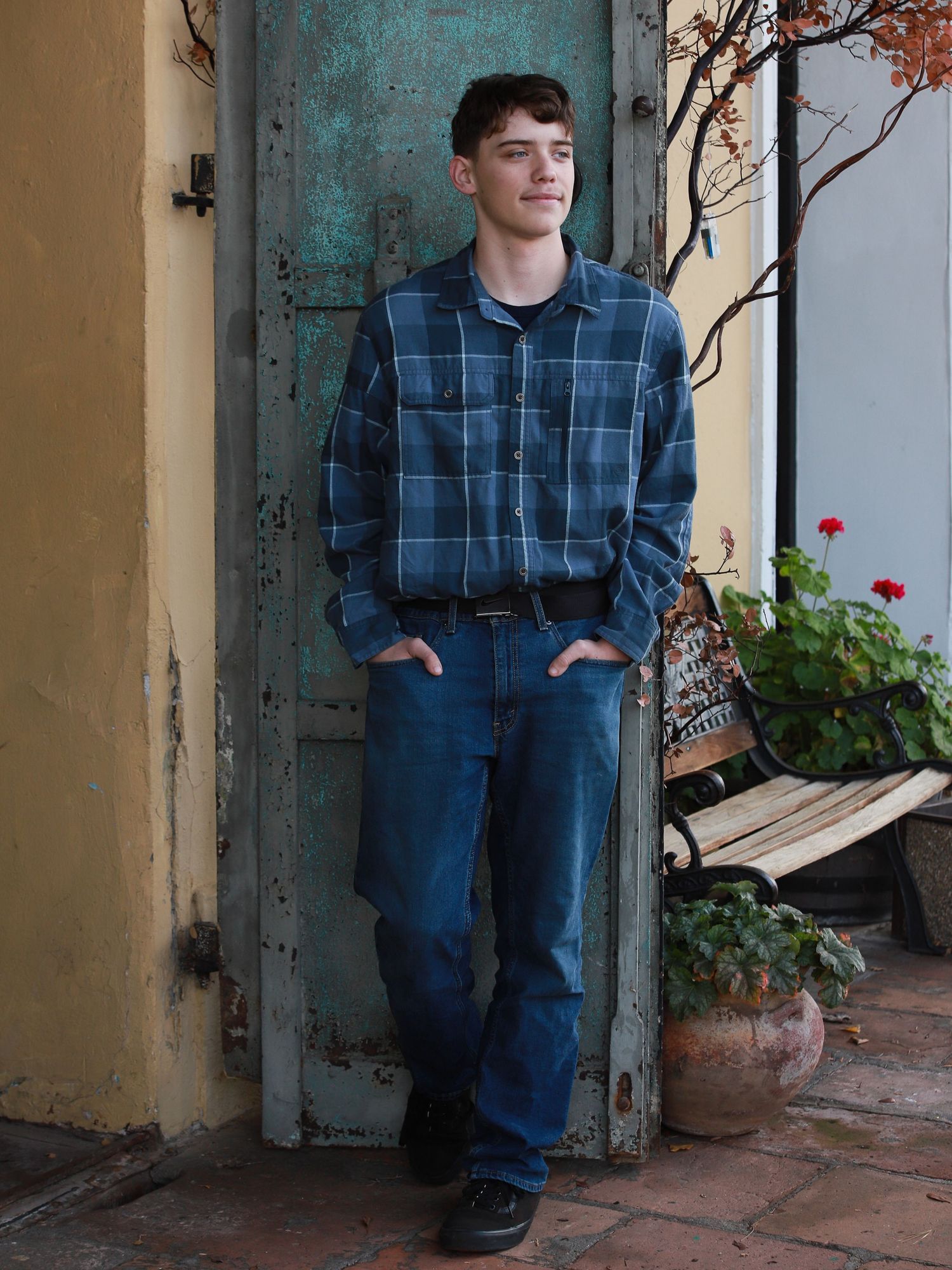 A person wearing a blue plaid jumpsuit sits casually on a porch with terracotta tiles and decorative branches.