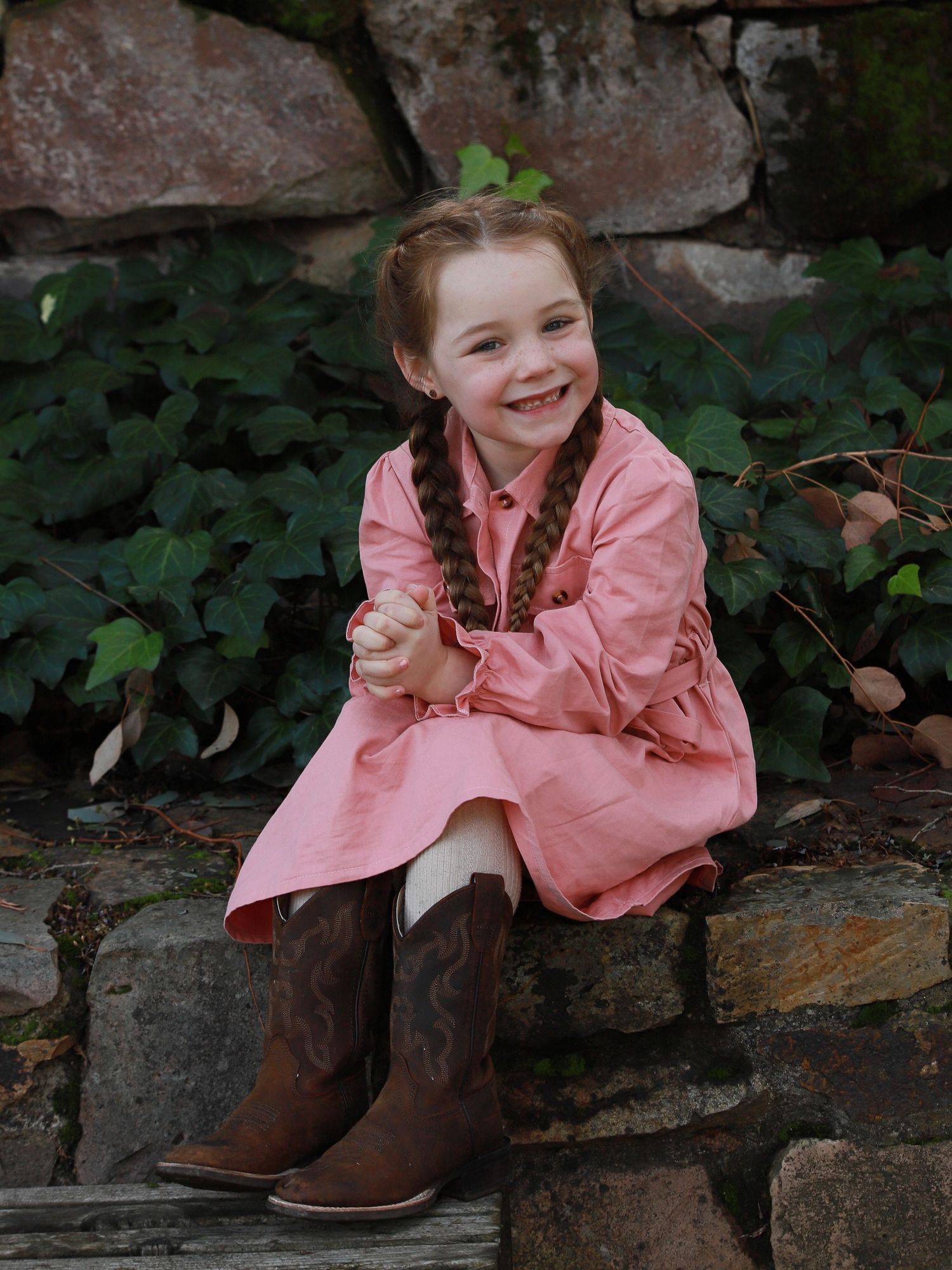 A child in a pink jacket sits on stone steps surrounded by green foliage in a garden setting.