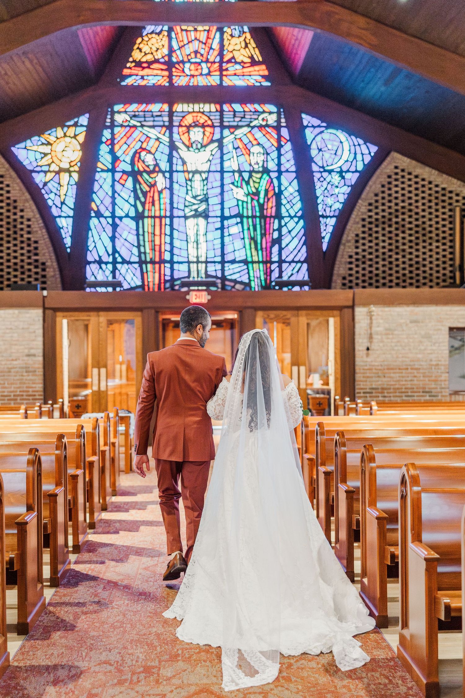 A couple stands in a church aisle beneath a stunning stained glass window with wooden pews and brick walls.