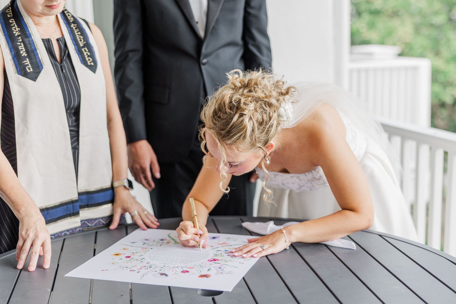 Someone signs a document on a gray table while others watch during a wedding ceremony.