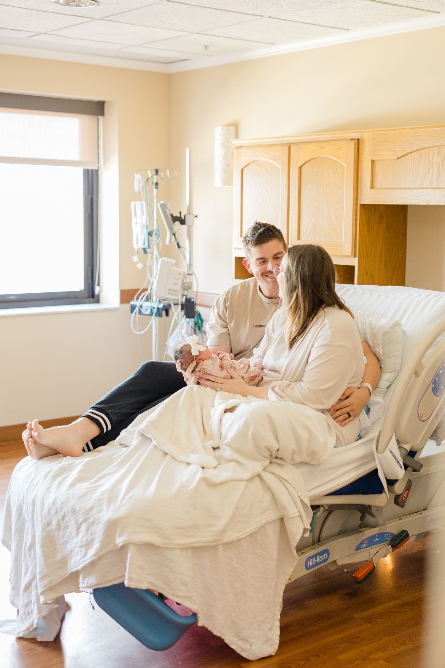 A couple lies together in bed under white sheets in a hospital room during Fresh 48 session in Pittsburgh, PA.