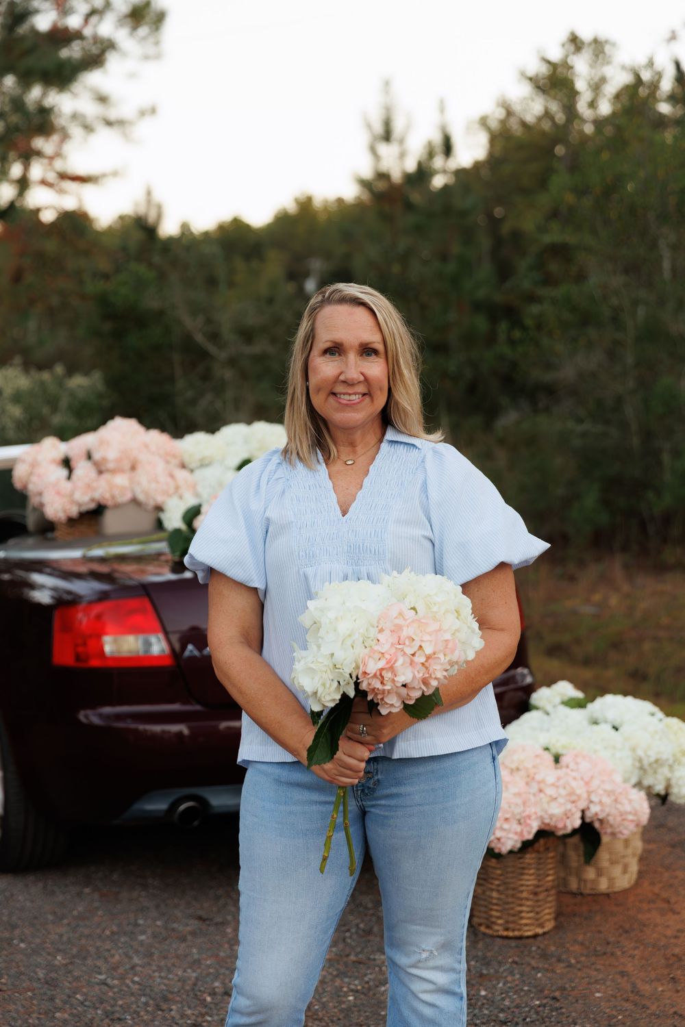 Woman in light blue shirt and jeans holds fresh pink and white floral bouquets against wooded outdoor backdrop.