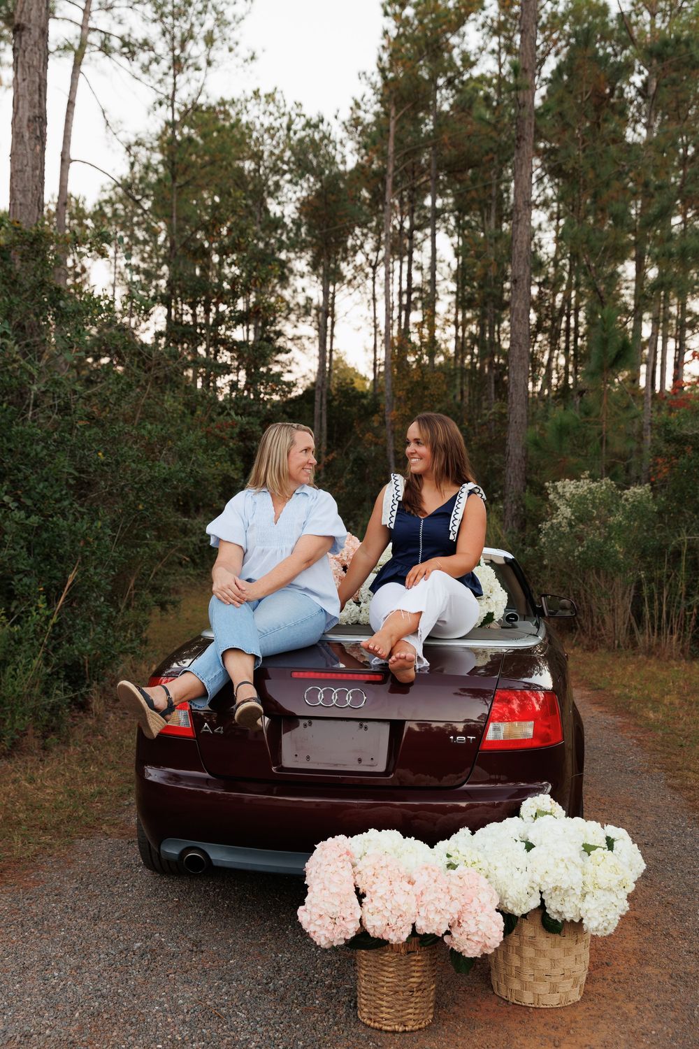 Two friends sit on the trunk of a burgundy Audi car with hydrangea flower baskets on a forest path at sunset.