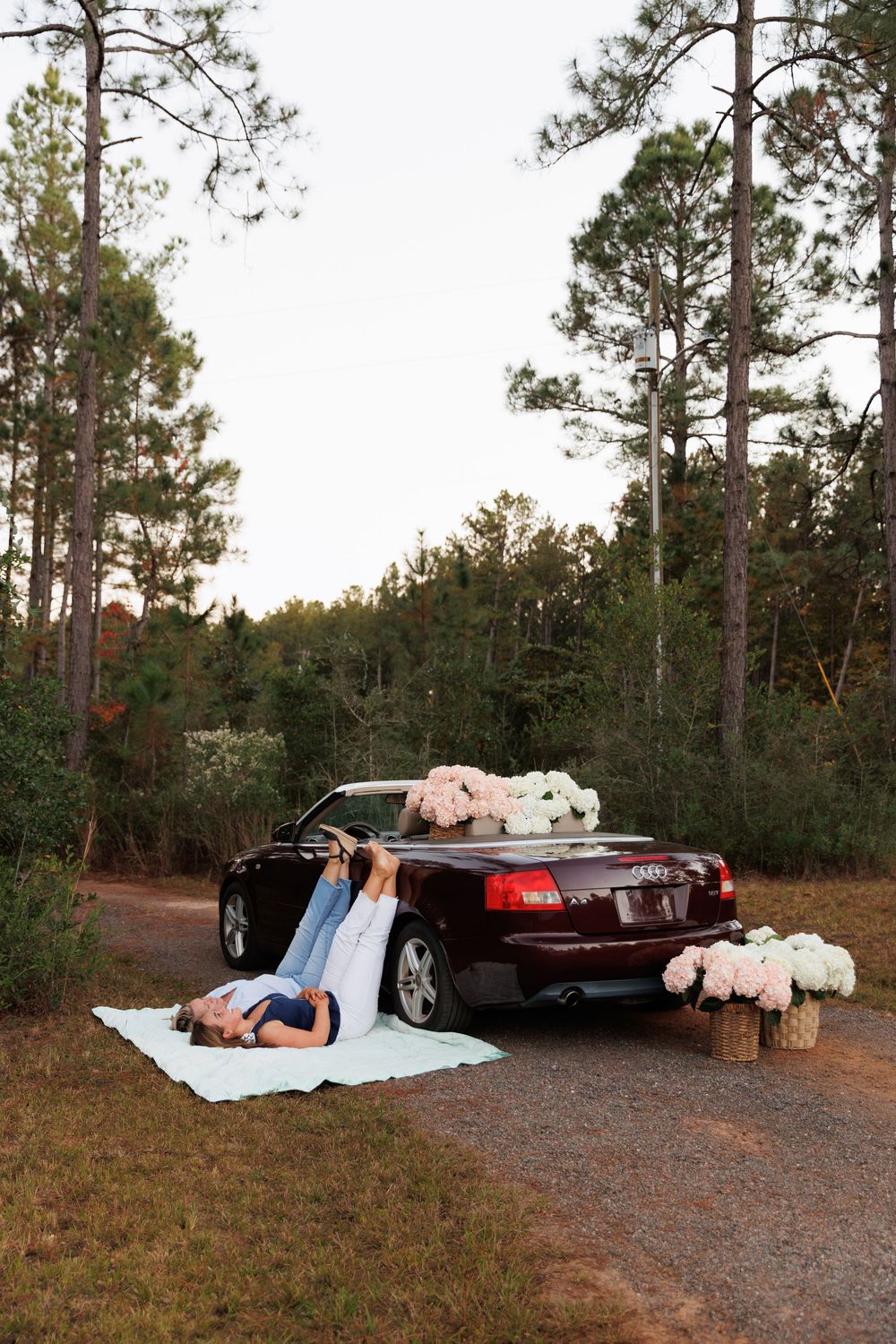 A couple lies on a blanket beside a car decorated with white flowers in a wooded outdoor setting at sunset.