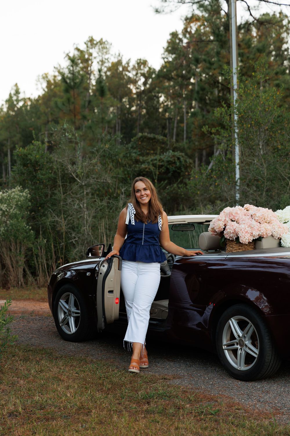 A woman in a navy top and white skirt poses with a black luxury convertible car on a wooded road.