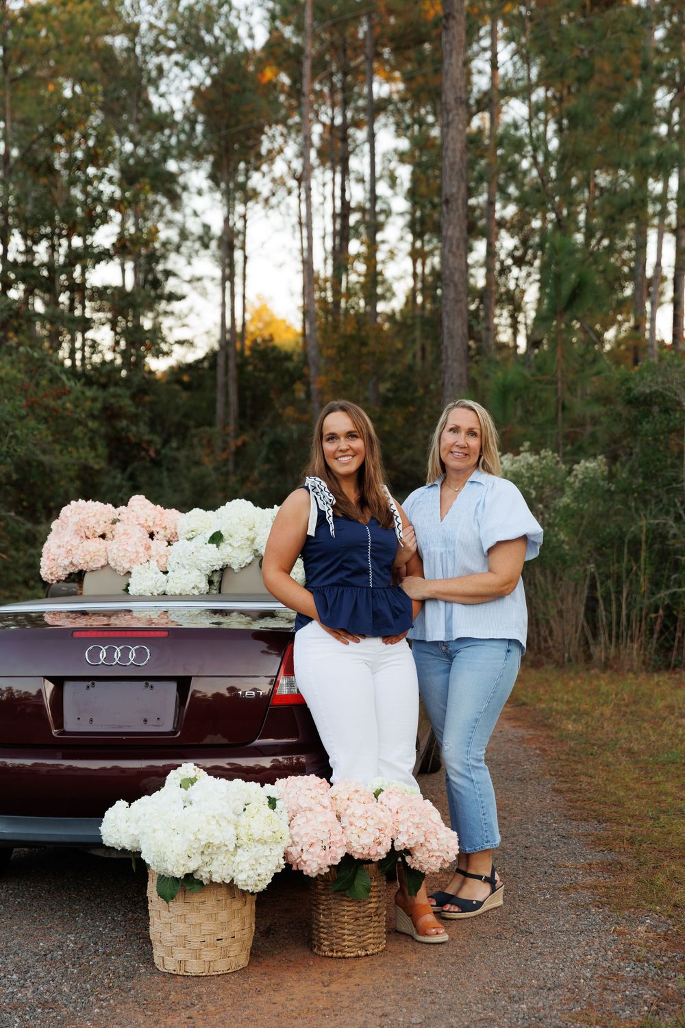 Two people stand beside an Audi car with pink and white hydrangeas in rustic baskets on a gravel path at sunset.