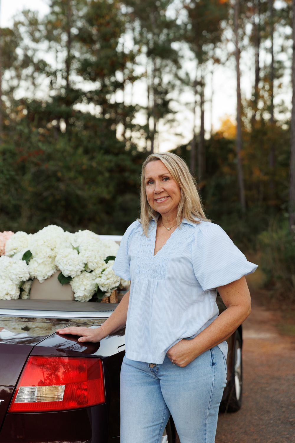 A woman in a white top and jeans poses casually near a car with white hydrangeas in a rustic outdoor setting.