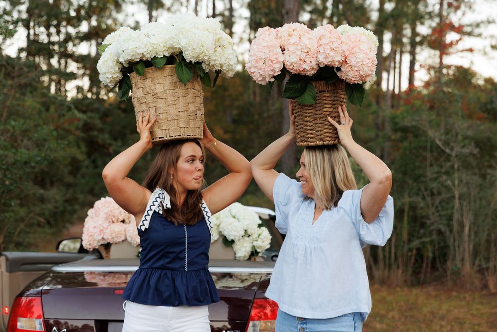 Two people hold woven baskets filled with pink and white flowers above their heads while standing near a truck.