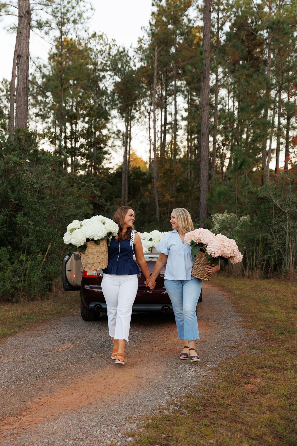 Two people walking down a dirt path in a pine forest while carrying bouquets of white flowers.