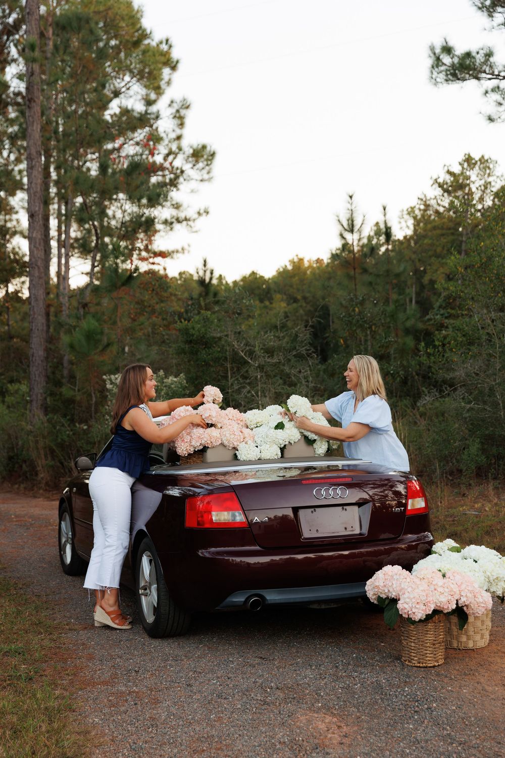 Two people loading hydrangea flowers into a black Audi convertible on a dirt road surrounded by pine trees at sunset.