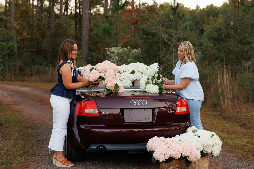 Two people loading white floral arrangements into the trunk of a burgundy Audi convertible on a wooded dirt road.