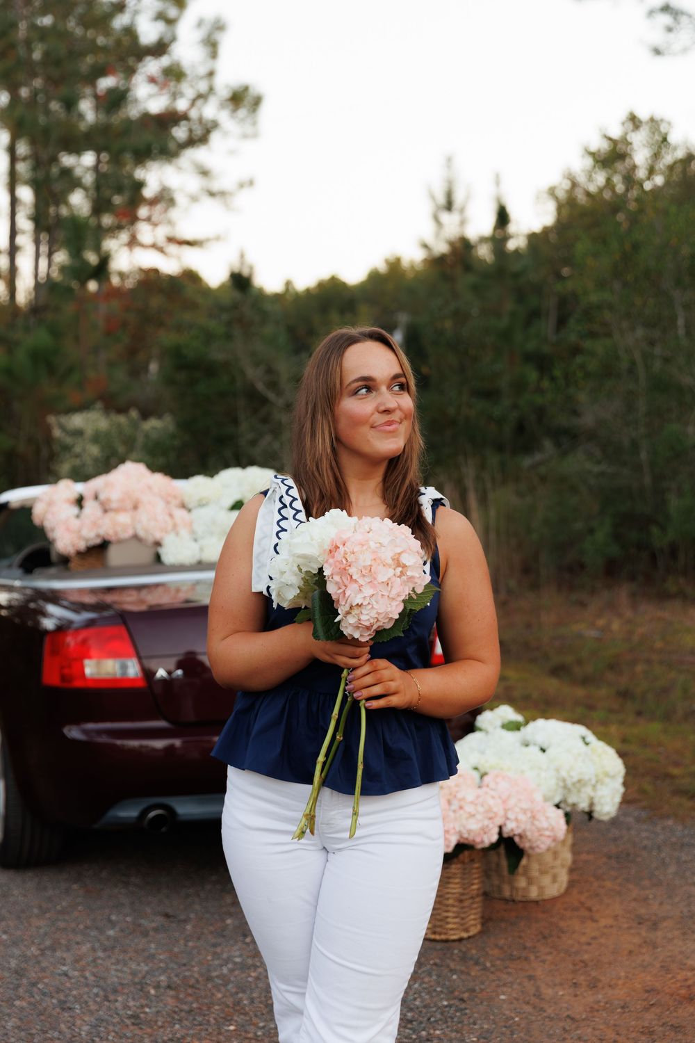 A woman in white pants and navy top holds pink flowers by a car with floral arrangements in baskets nearby.