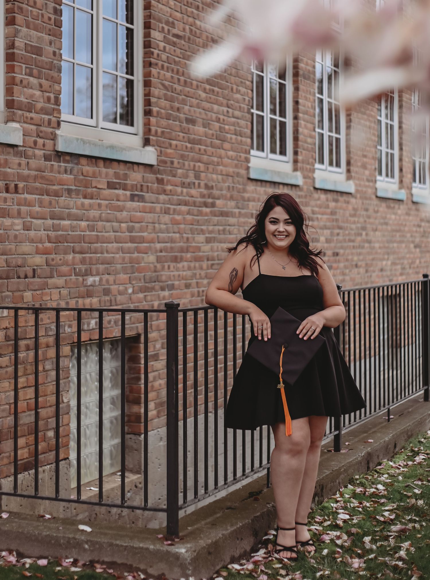 A woman in a black dress poses confidently against a brick building with white windows.