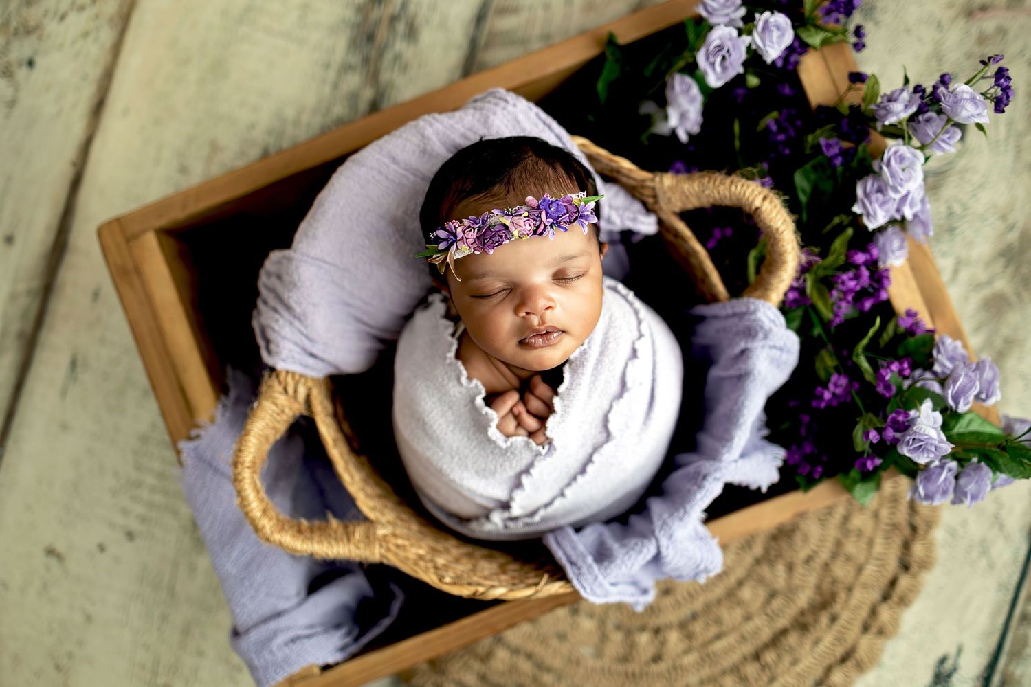 A sleeping baby in a yellow crib wearing a purple floral crown next to fresh purple flowers.