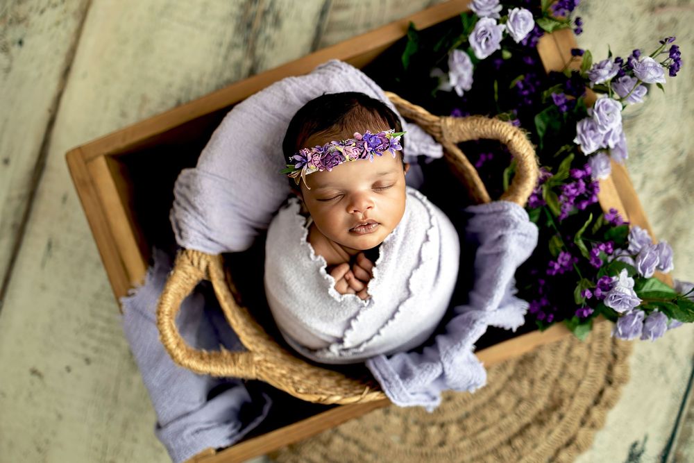A sleeping baby in a yellow crib wearing a purple floral crown next to fresh purple flowers.