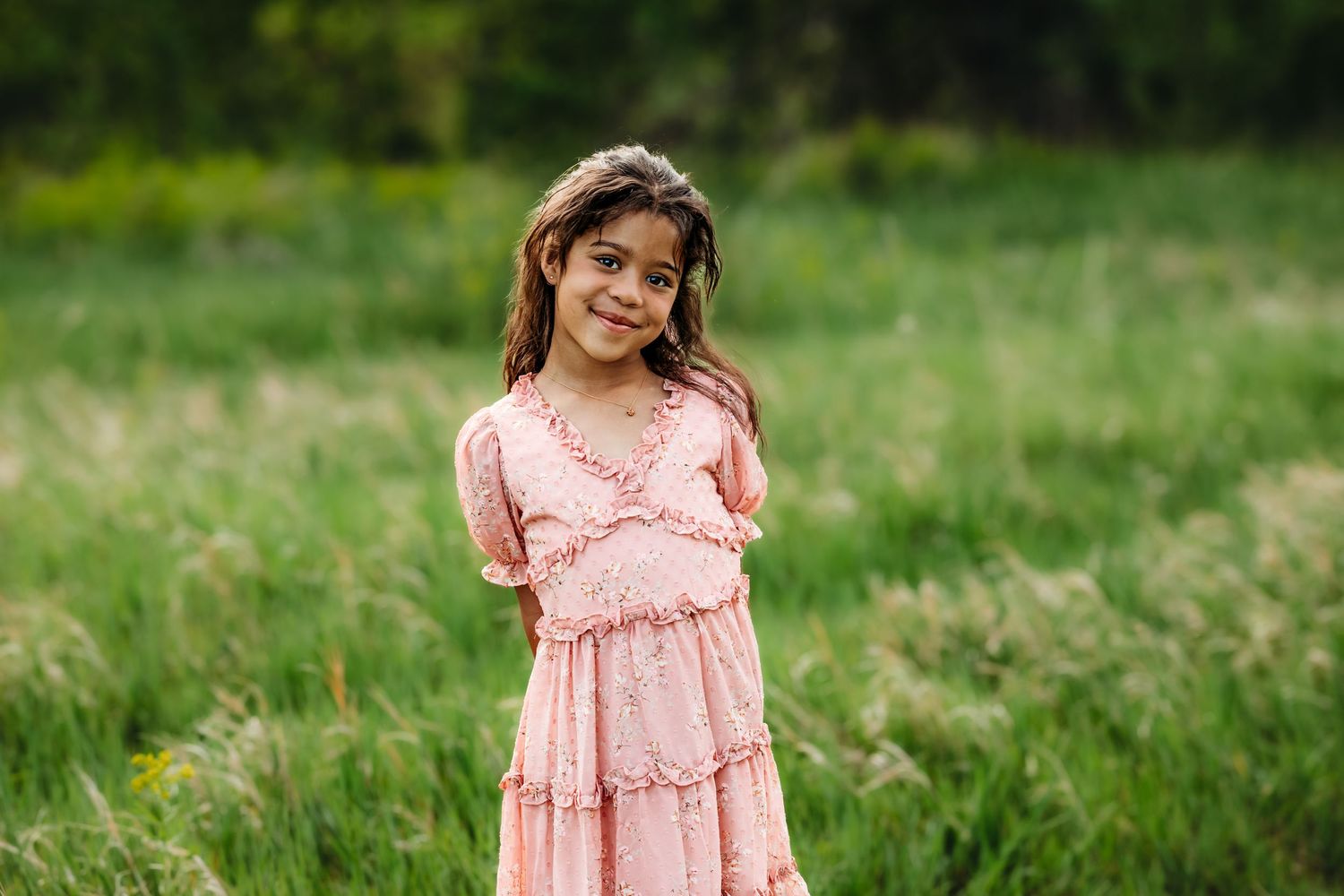 A child wearing a pink ruffle dress poses in a sunny green field with wildflowers.