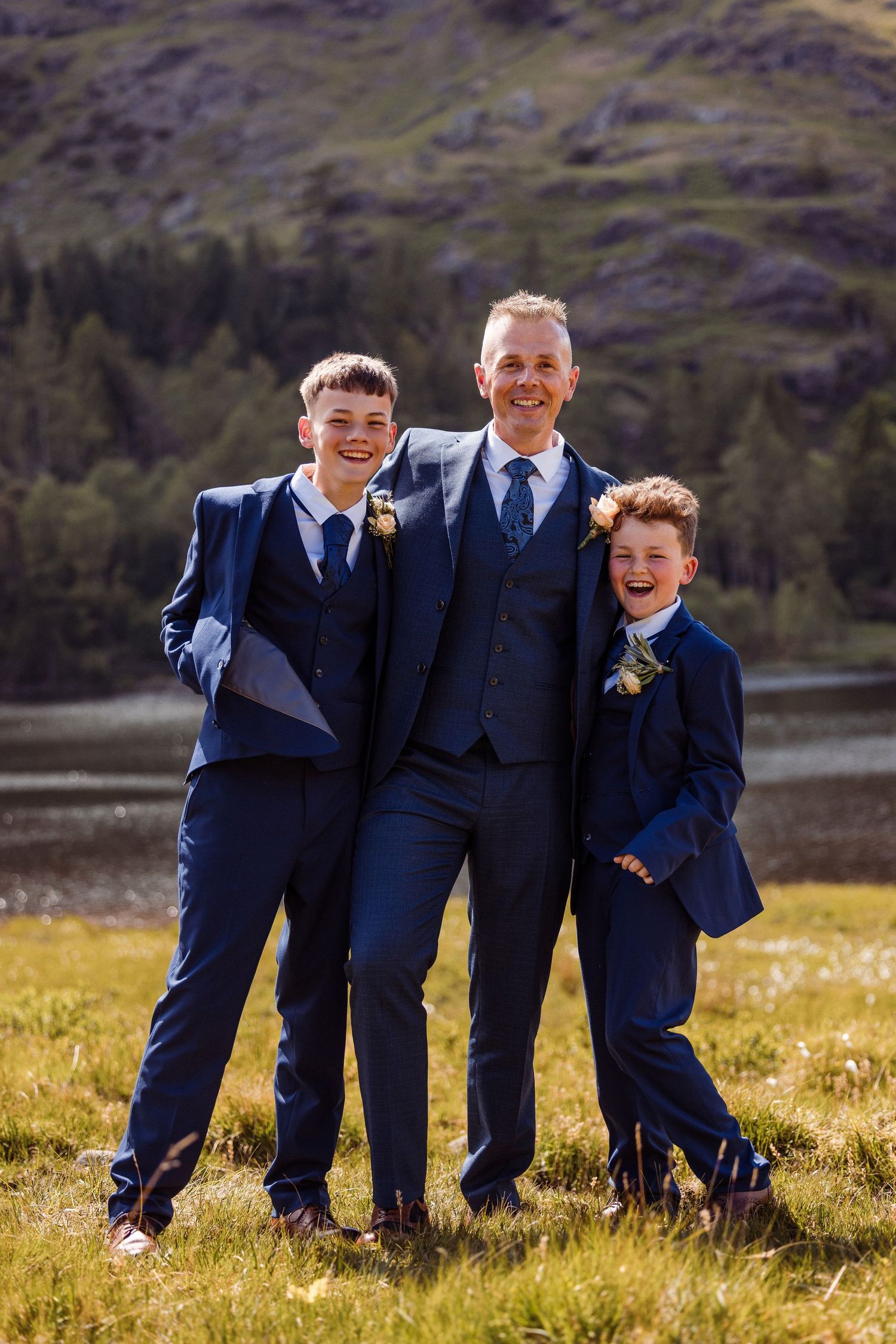 Three people in matching navy blue suits and boutonnieres pose together on a grassy hillside with mountains in background.