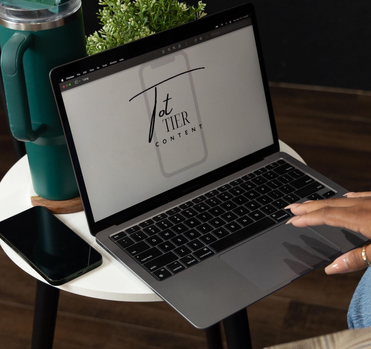 Laptop displaying a logo design on screen with a teal water bottle and smartphone on wooden table.