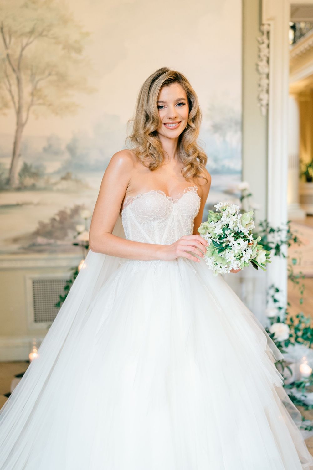 A bride in a white ballgown wedding dress poses with a white and green bouquet in an elegant room with wall murals.