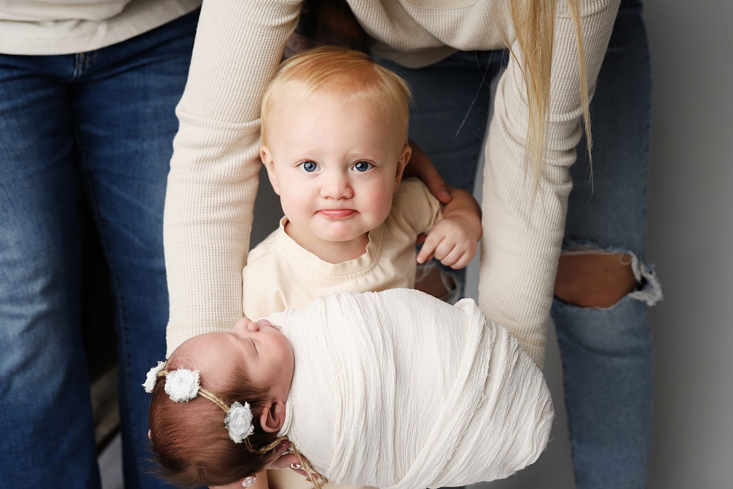 A small child in a cream-colored outfit stands between two adults while holding a wrapped bundle.