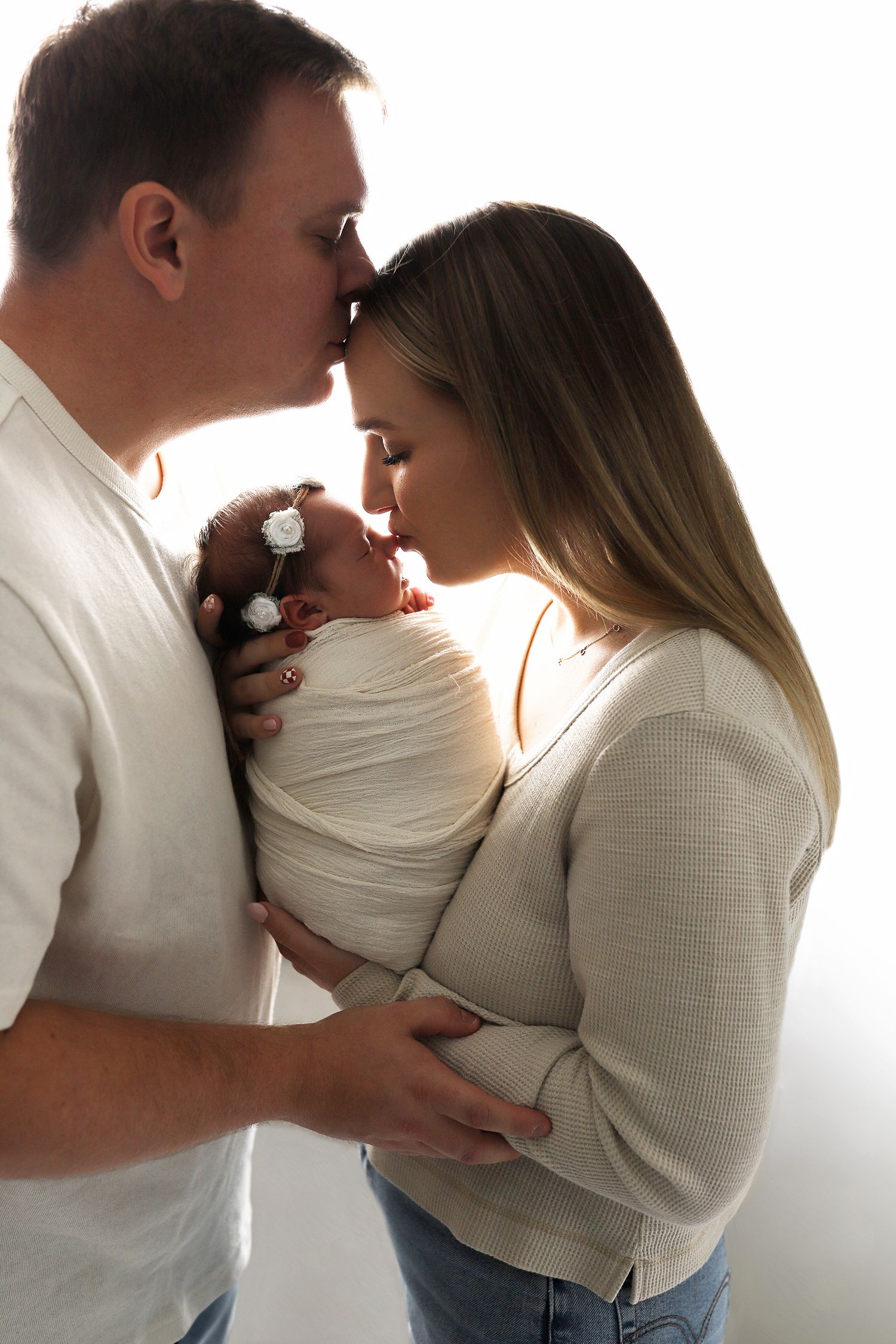 Intimate family portrait series showing parents embracing their newborn baby in soft white clothing against neutral backdrop.