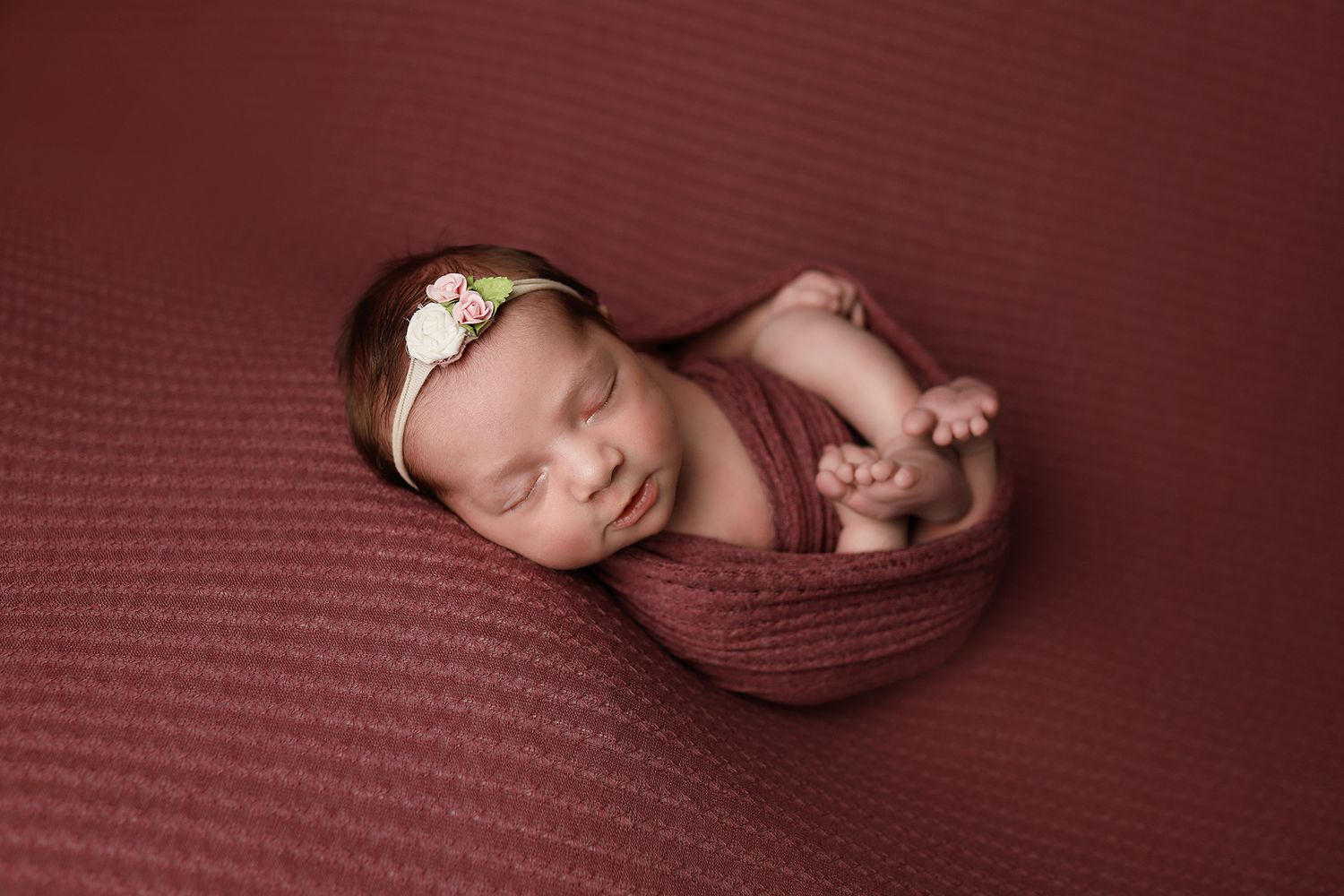 Sleeping newborn baby curled up on soft burgundy textured blanket with matching wrap and headband.