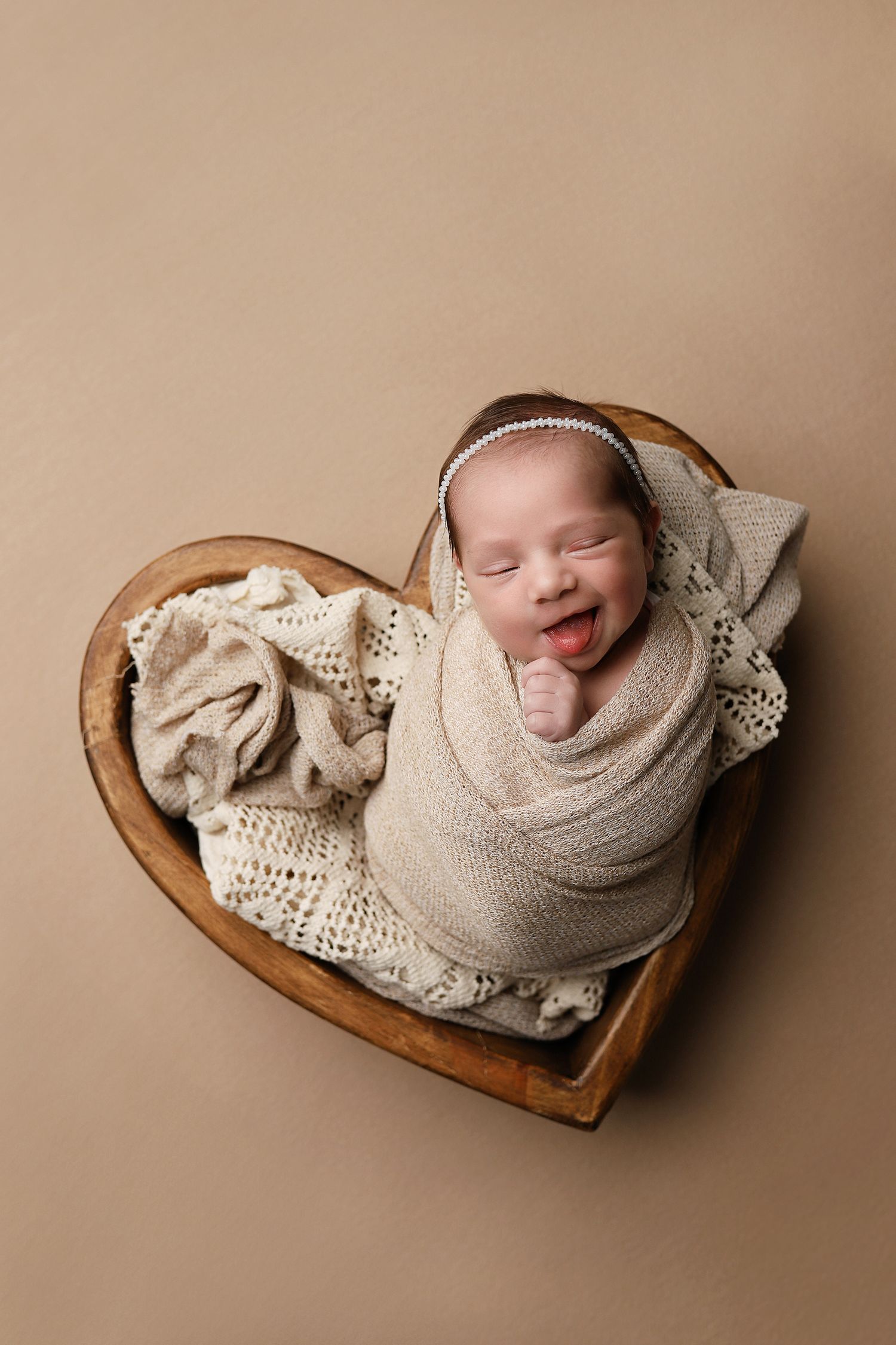 Sleeping newborn wrapped in beige swaddle lying in heart-shaped wooden bowl on neutral backdrop.