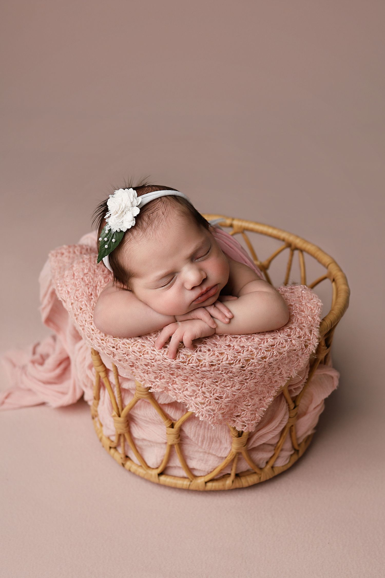 Newborn sleeping peacefully in pink lace-lined basket with floral headband on mauve background.