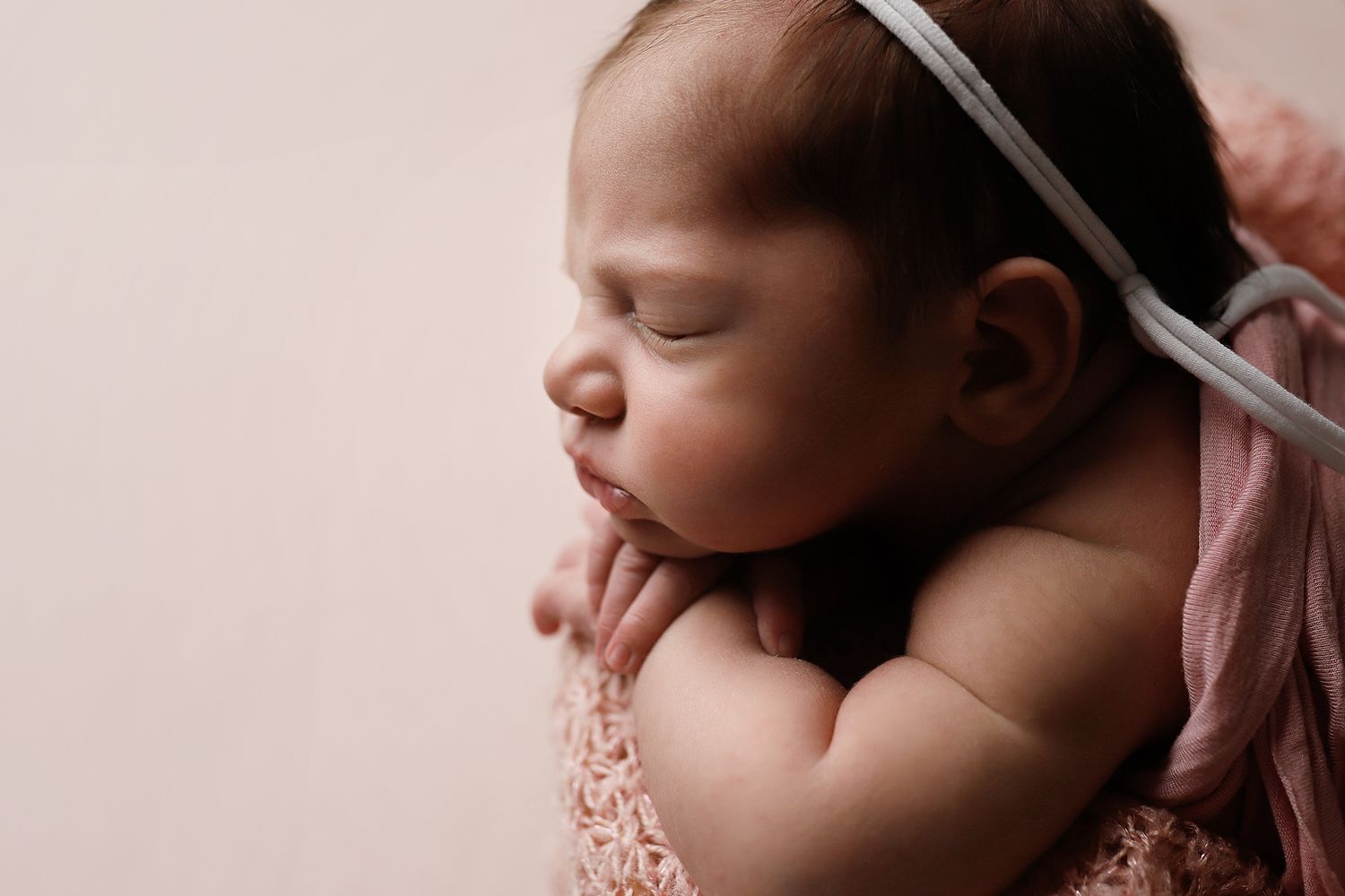 Sleeping newborn baby wearing a delicate headband rests peacefully against a soft pink backdrop. Santaquin in-home newborn photo session.