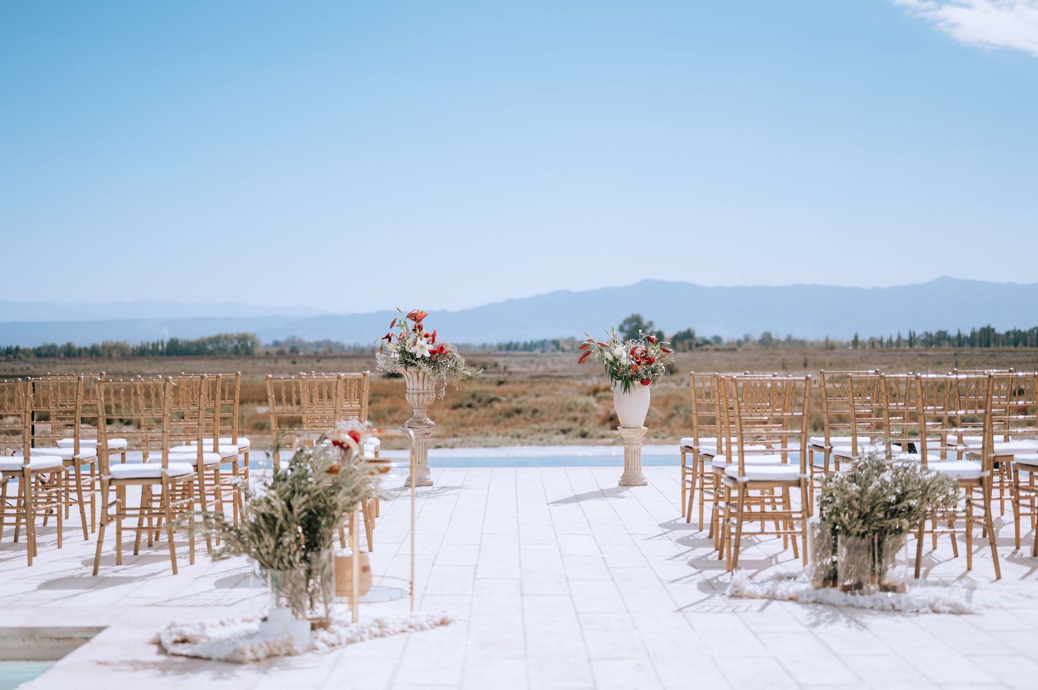 Elegant outdoor wedding ceremony setup with floral arrangements, wooden chairs, and mountain views under blue sky.
