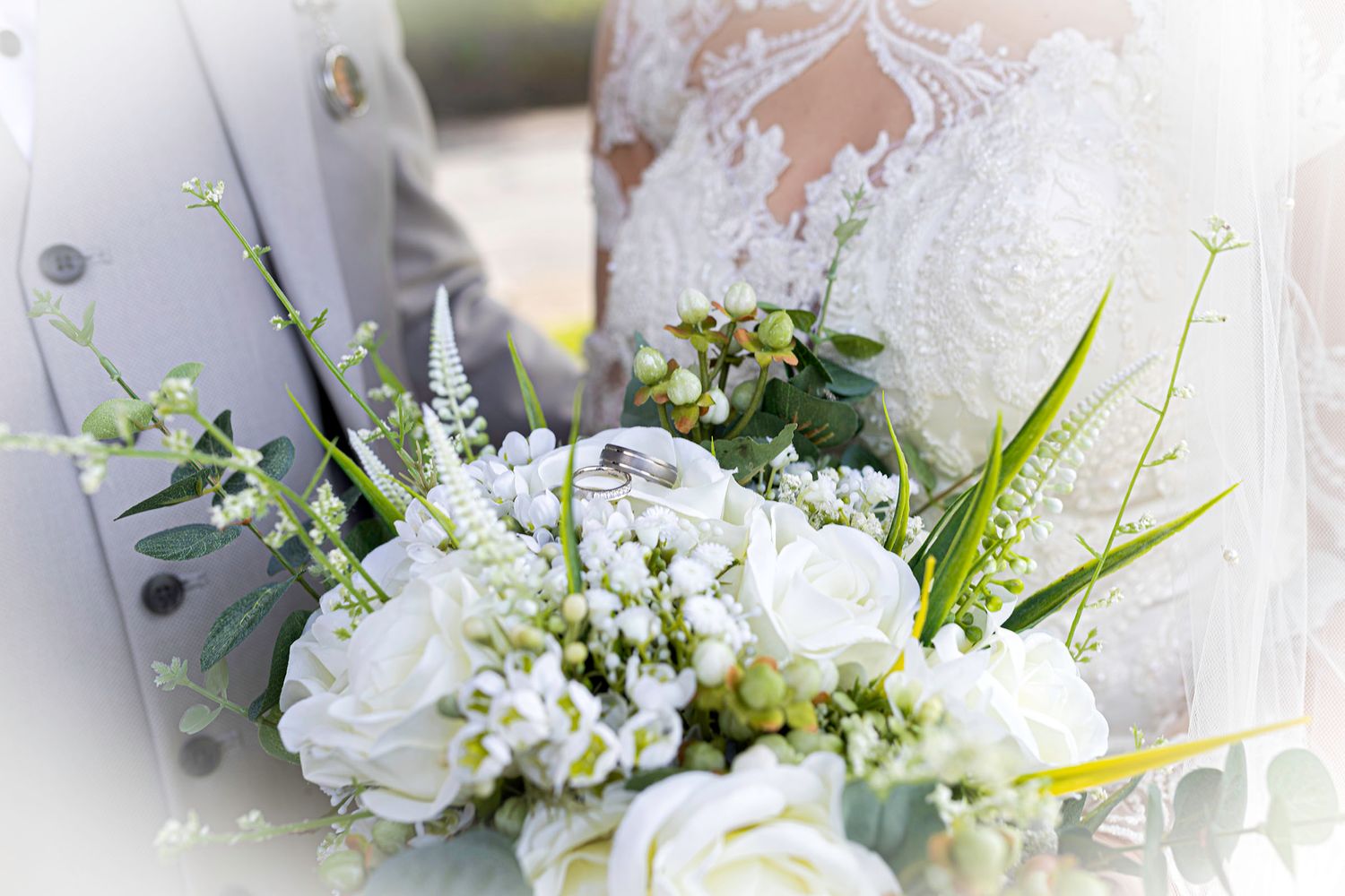 Wedding bouquet of white roses and greenery with two gold rings resting on top, held by bride in lace gown. heritage park south Wales