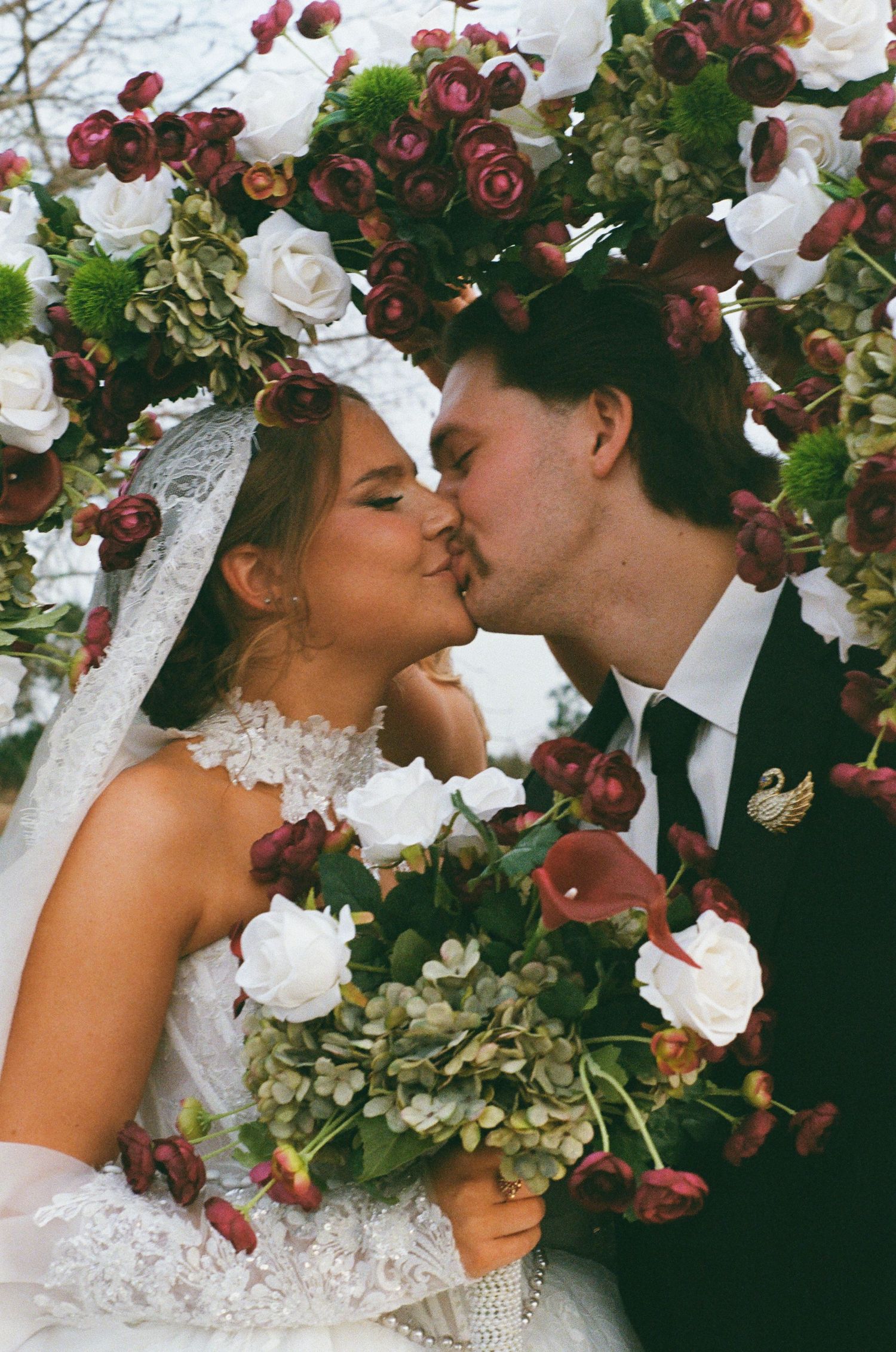 Bride and groom kissing under floral arch with white and burgundy roses, bride holding bouquet, wearing lace veil.
