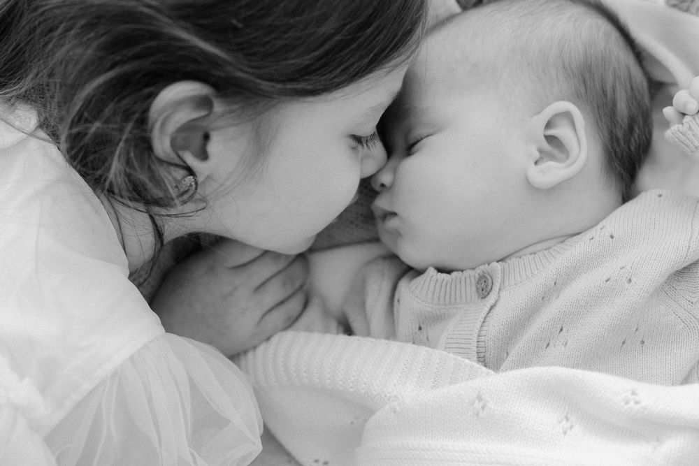 Black and white photo of a young girl tenderly touching noses with a sleeping newborn baby sibling.