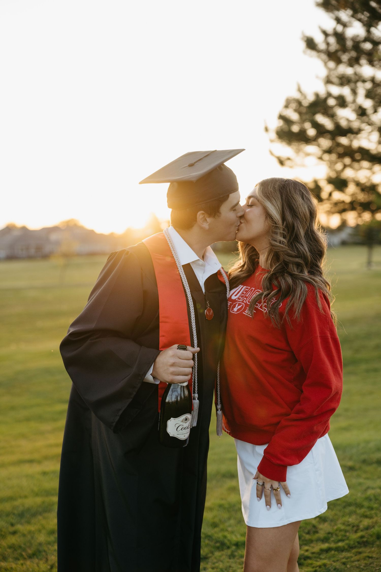 A couple shares a kiss at sunset, one wearing a graduation cap and gown, the other in a red sweatshirt and white skirt.