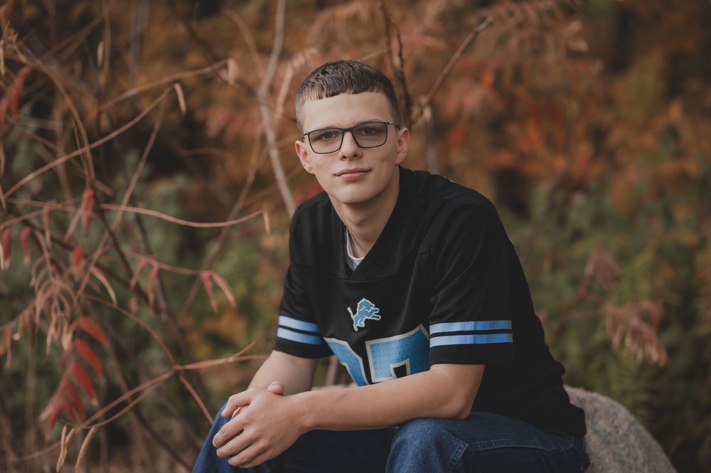 Teen boy in Detroit Lions jersey and glasses sits outdoors among autumn foliage for senior portrait.