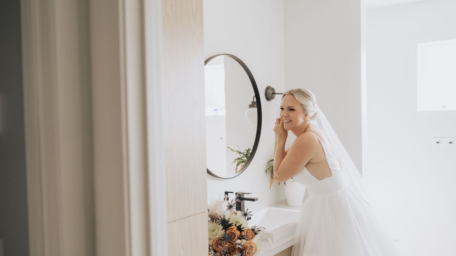 Mariée souriante en robe et voile devant un miroir de salle de bain – préparation mariage élégante et photo naturelle.