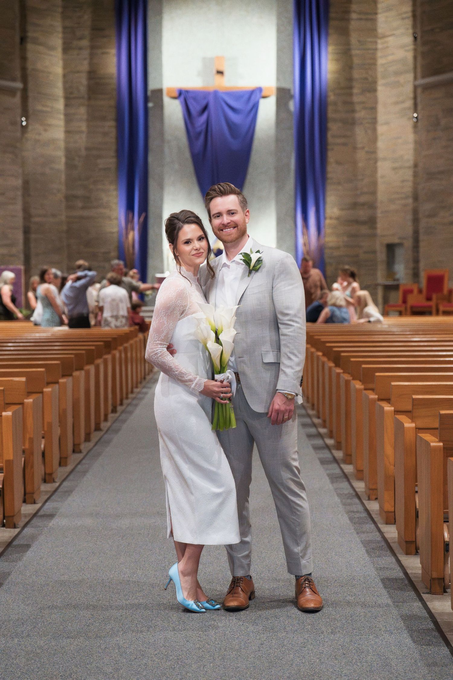 Wedding couple posing in church aisle with wooden pews and blue drapes in background.