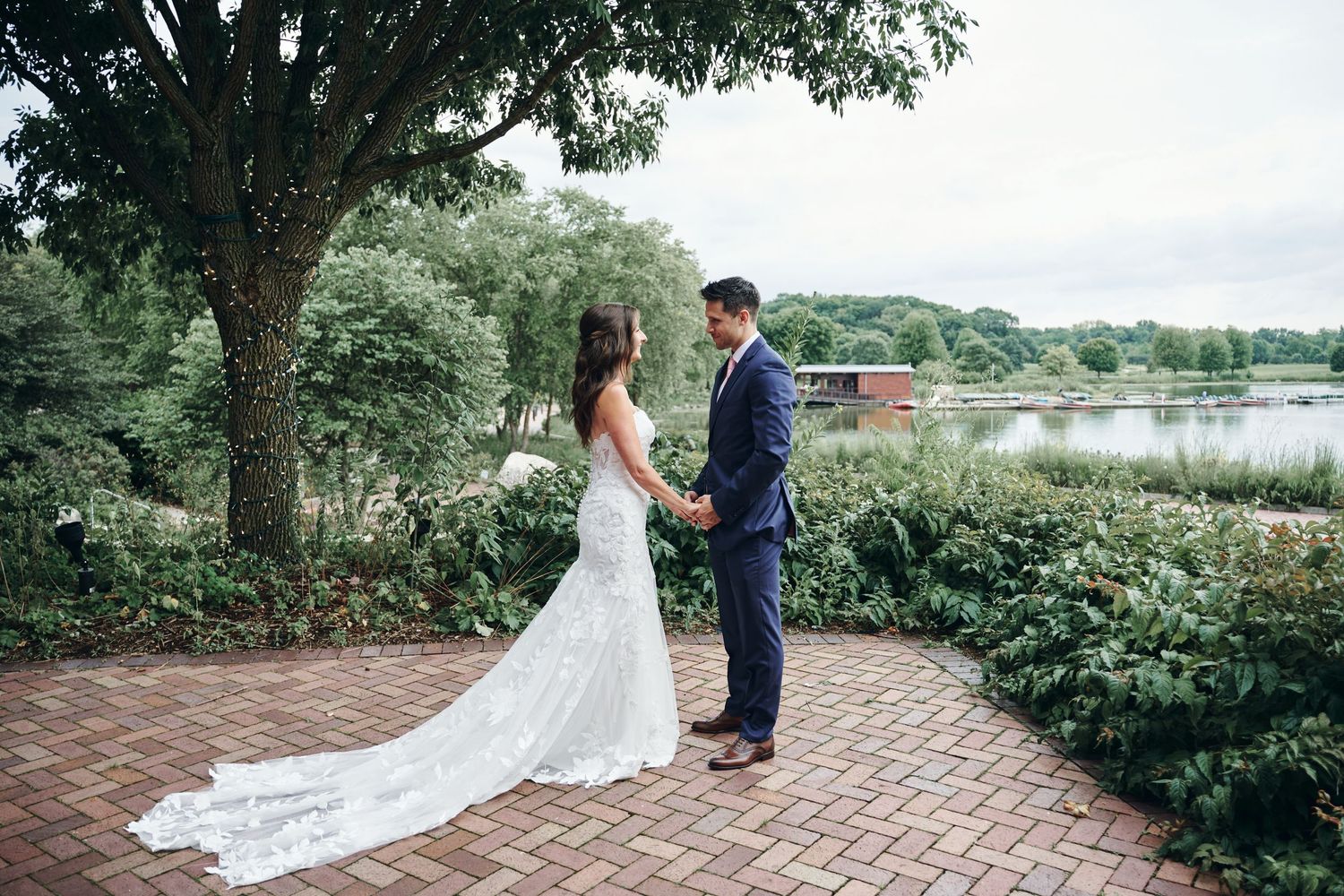 A couple in wedding attire stands on a brick pathway beside a lake with lush greenery and trees in the background.