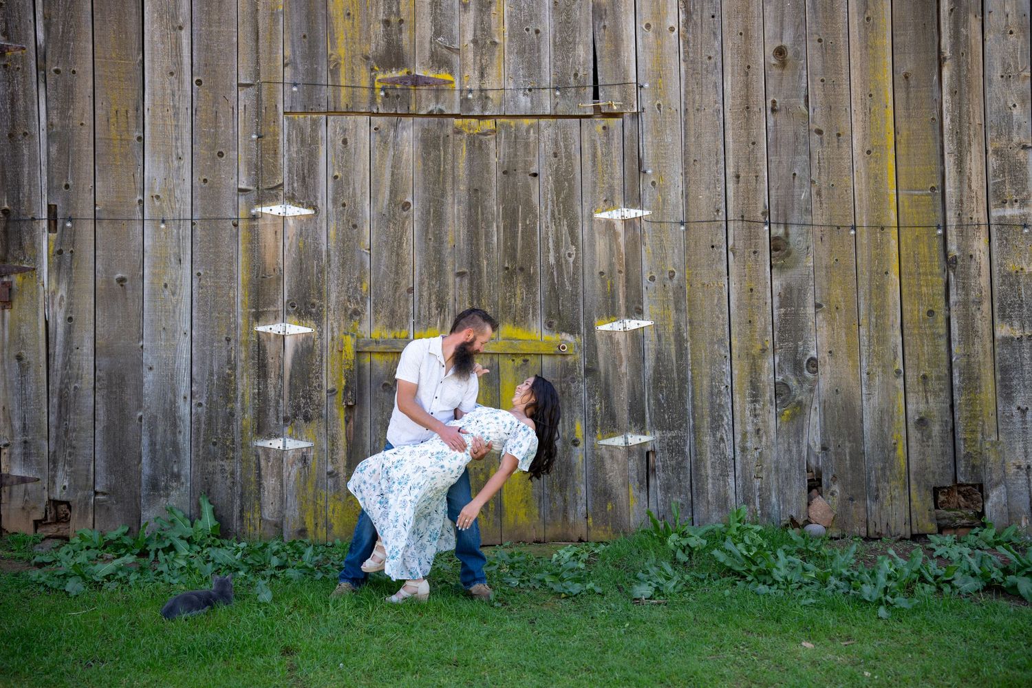 Two people share a romantic dip pose against a rustic wooden wall with grass and white lights in the background.