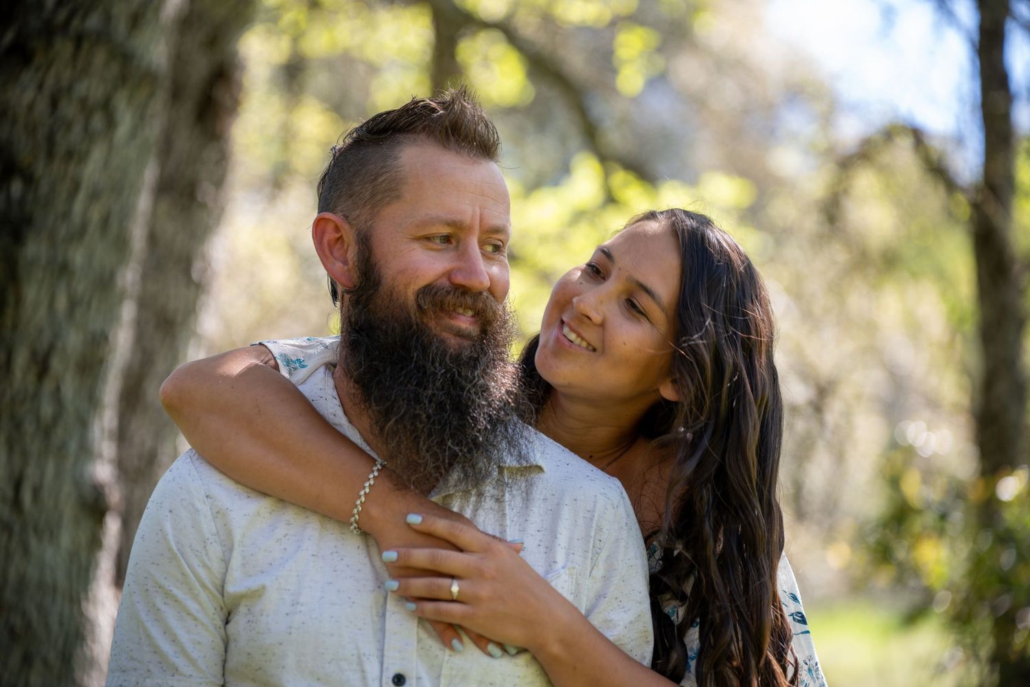 A couple shares intimate moments outdoors while surrounded by sunlit trees and natural scenery.