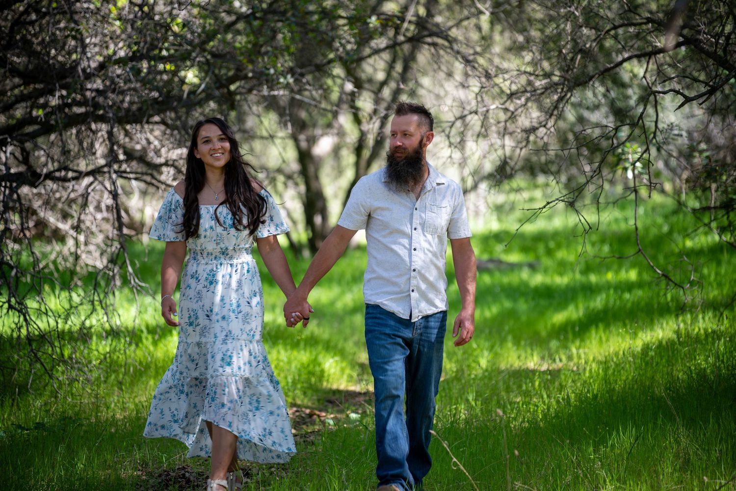 A couple holds hands while walking through a sunlit spring orchard with blooming trees in the background.