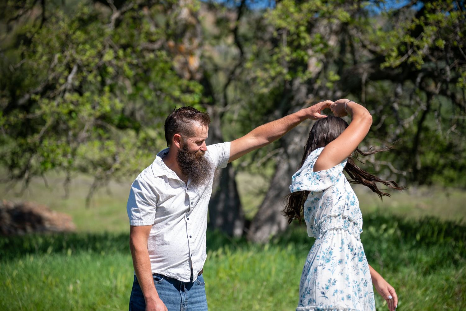 A couple dances together in a grassy meadow surrounded by trees on a sunny day.