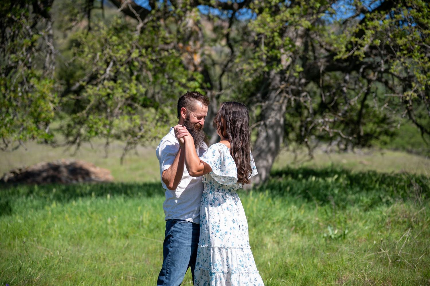 Two people in casual clothing stand together in a sunny meadow with green trees and foliage in the background.