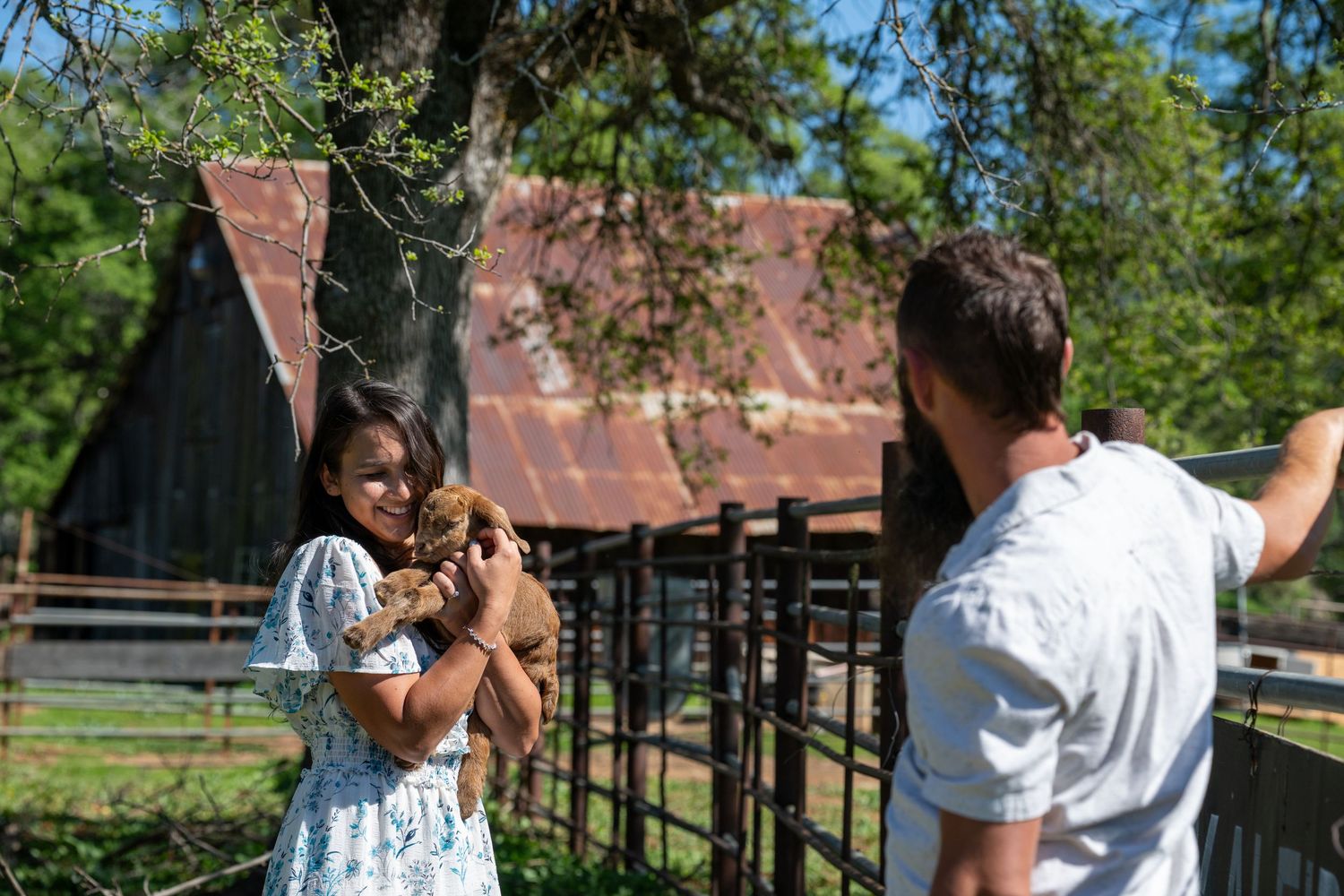 A couple shares a romantic moment outdoors at a rustic venue with wooden buildings and green trees in the background.