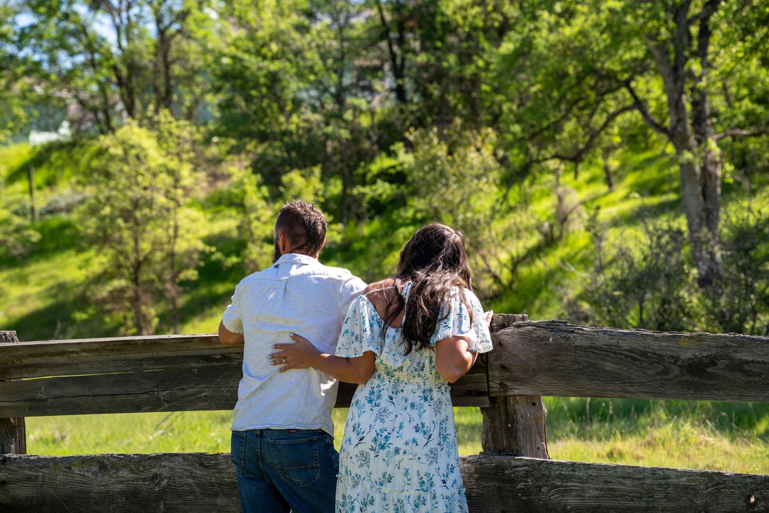 A couple in summer attire leans against a wooden fence overlooking a sunny green hillside with trees.