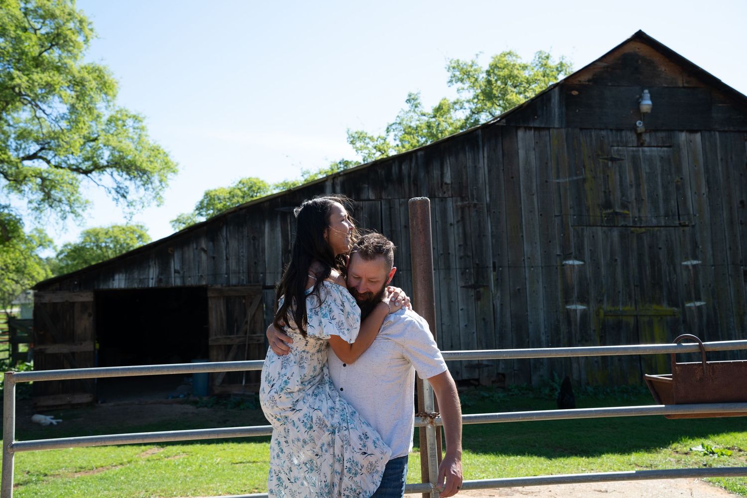 A romantic couple shares a tender moment in front of a rustic black barn on a sunny day.