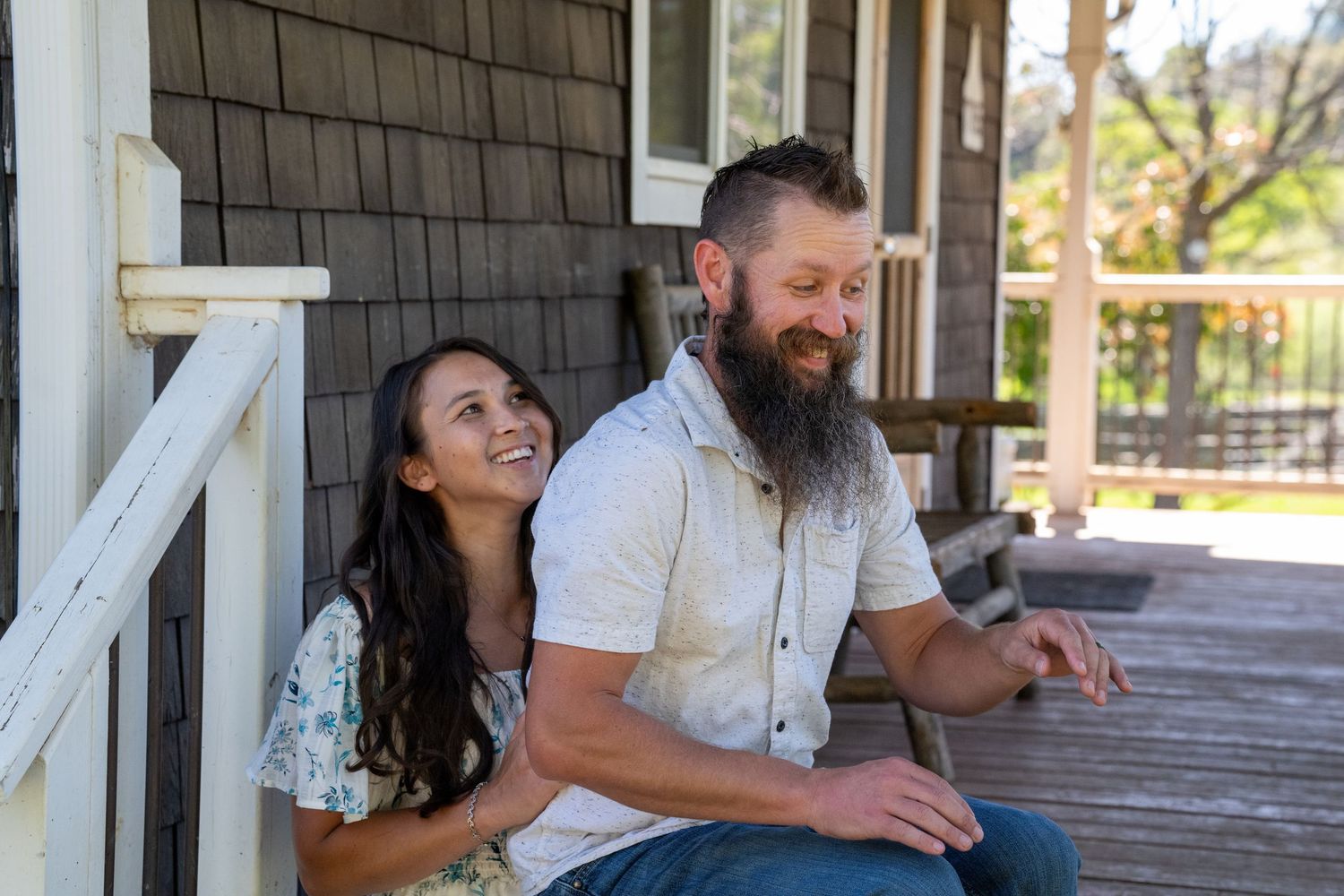 A couple sits and laughs together on a rustic wooden porch during a sunny afternoon.