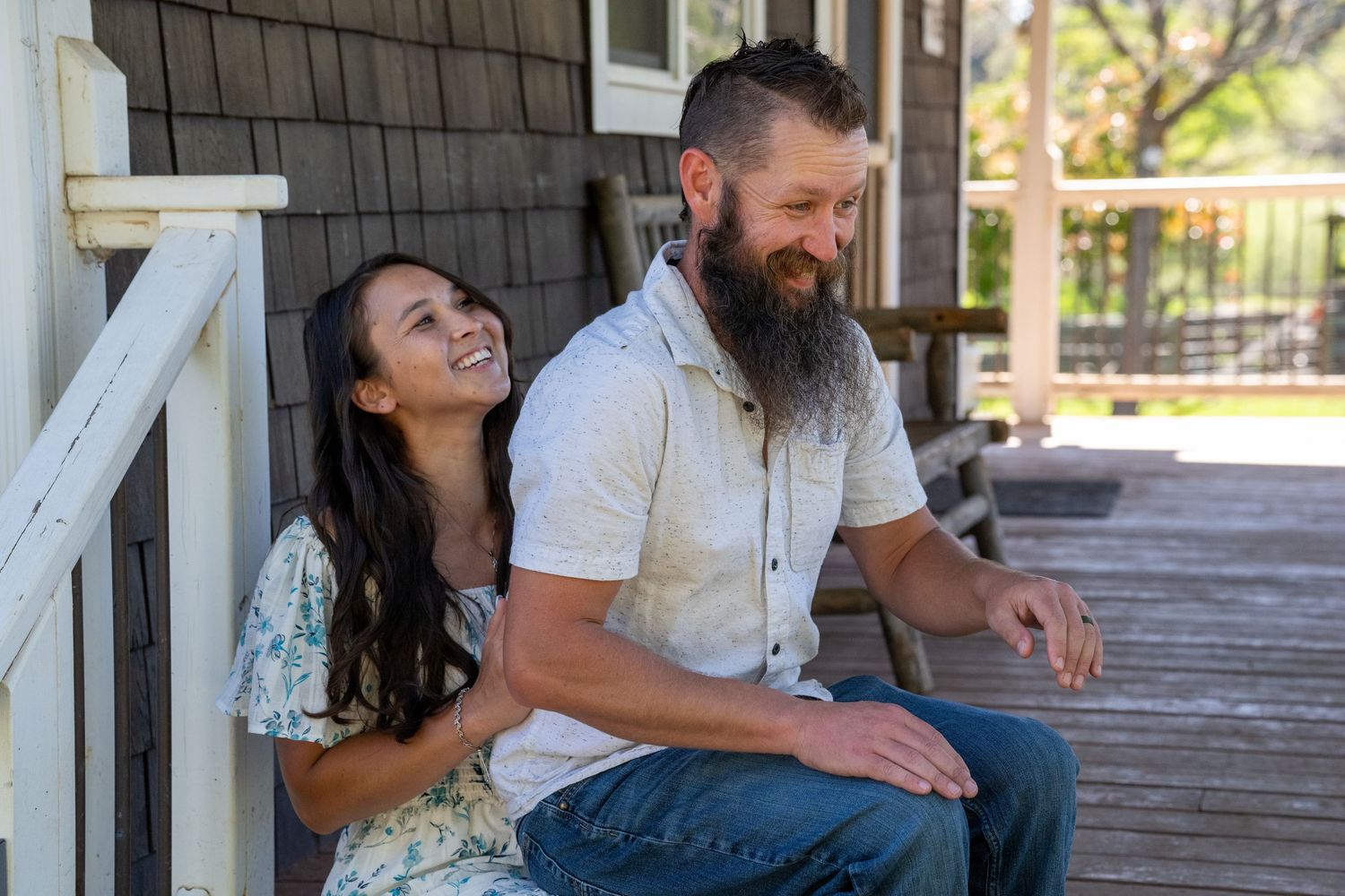 A couple sits together and shares a laugh on a wooden deck in casual white clothing.