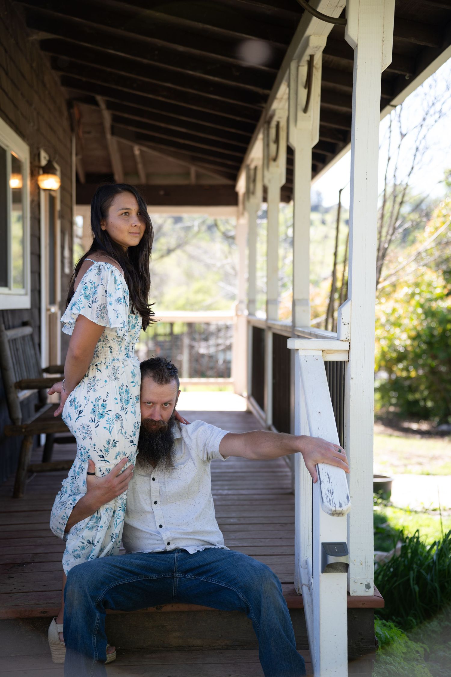 A couple shares a playful moment together on a rustic covered porch with white pillars and natural lighting.